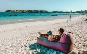 A man sitting on a bean bag at the beach, laptop in hand