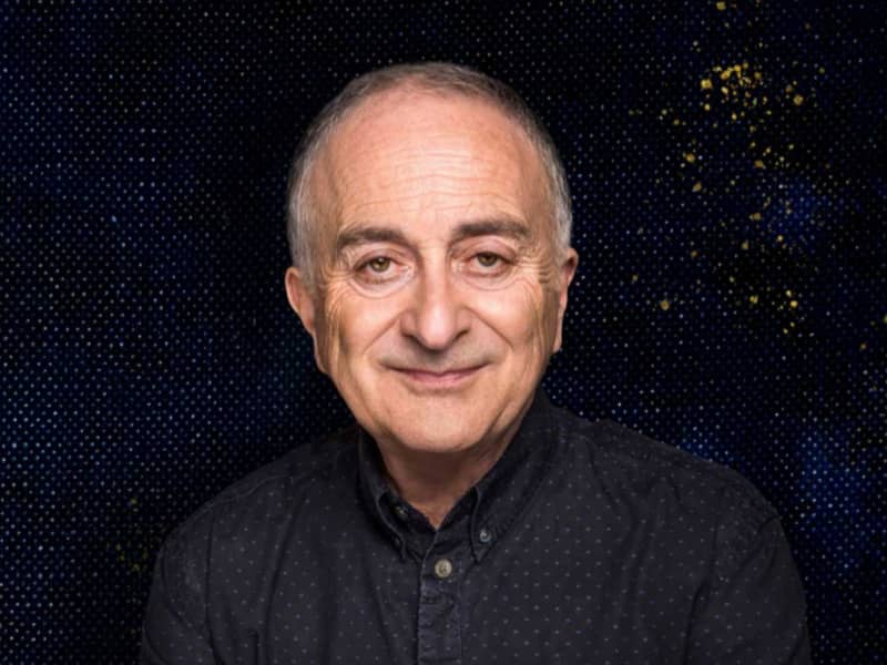 Sir Tony Robinson is a slightly smiling bald man wearing a black shirt with a black background behind him.