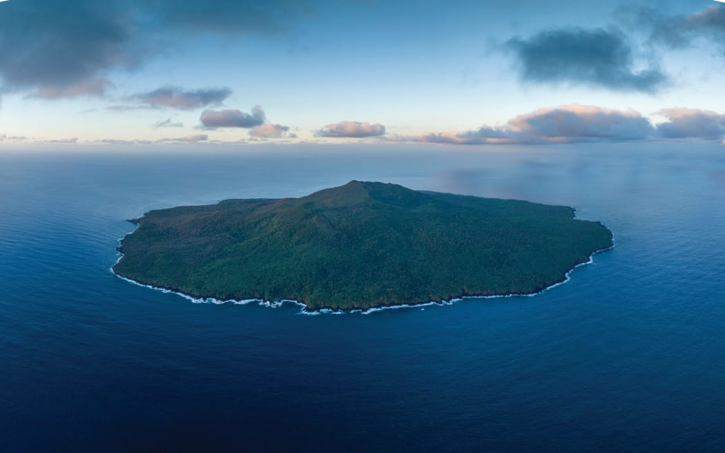 An aerial image of a circular island with a mountain/volcano at its centre. The island is covered in green vegetation and sits in a blue expanse of ocean beneath a cloud-studded sky.