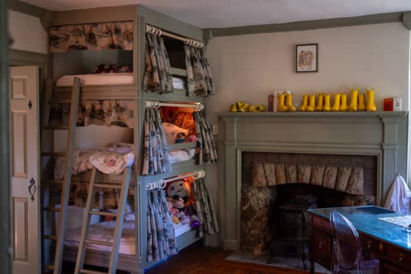 A kid's bedroom with three bunker beds on top of each other and traditional-style curtains covering each. Small yellow boots sit on top of the ledge of the fireplace near the beds.