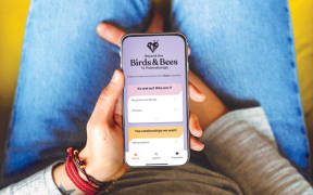 A young person holds a phone with the screen showing the Te Puāwaitanga: Beyond the Birds & Bees app