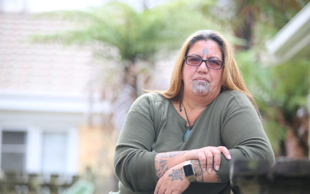 Associate Professor Leonie Pihama sits outside. She is wearing a dark green top with a pounamu toanga around her neck. She also wears glasses.