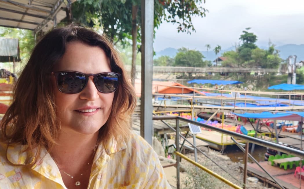 Jane Daniell sits by a riverside market in Laos. She is wearing sunglasses and a yellow patterned shirt.
