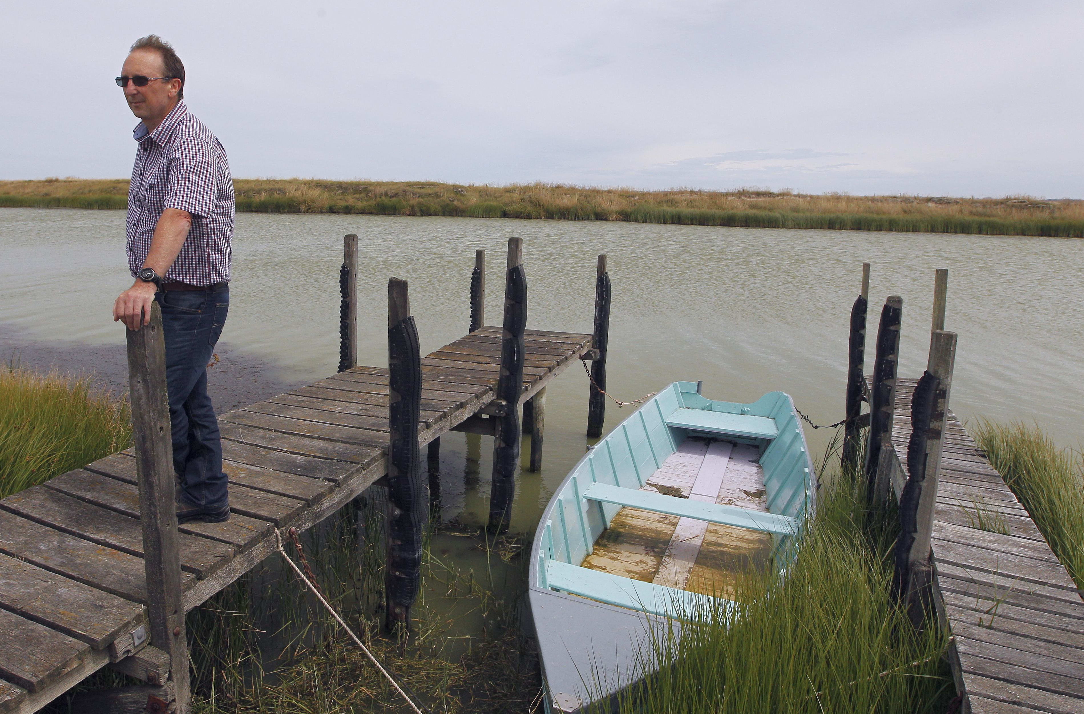 Man stands on small wooden jetty on edge of murky river by small wooden boat