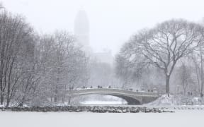 People walk across the Bow Bridge in a snow-covered Central Park in New York City on December 27, 2025. New York City receieved around 4 inches (10 centimeters) of snow overnight. Airlines canceled 1,500 US flights during the peak holiday travel period Friday, with severe winter storm warnings and heavy snow forecast across parts of the Midwest and northeast. (Photo by TIMOTHY A. CLARY / AFP)