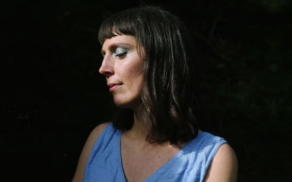 Amiria Grenell in blue dress, eyes closed in front of a black background.
