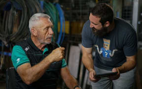 Luke and Mark are animatedly talking. They are in a warehouse with lots of equipment stacked in the background. Mark is seated while Luke bends down to show him something on an iPad.