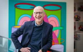 Dr Ezekiel Emanuel, a bald smiling man in a bule suit, leans with one hand on hip against a railing with a brightly coloured artwork behind him.
