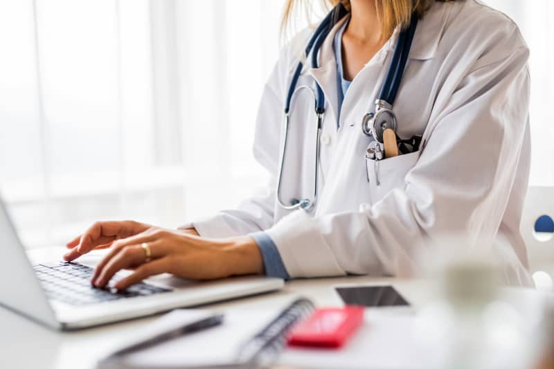A woman in a white coat and stethoscope on her shoulders types on a laptop.