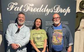 Two men with grey beards and wearing shirts, stand on either side of a dark-haired woman in a green t-shirt in front of a poster reading 'Fat Freddy's Drop'.