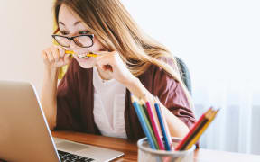 A girl wearing glasses grips a pencil between her hands and teeth as she looks at a laptop screen.