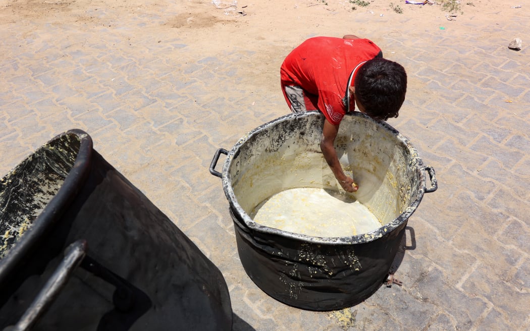 A Palestinian boy scrapes off bits of lentil soup remaining in a cooking pot, in front of a food distribution point at a displacement camp near Gaza City's port on 22 May, 2025.