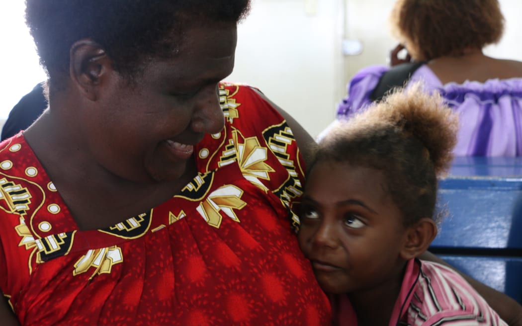 A woman in a red dress holds a younger girl in a hug. They smile at each other.