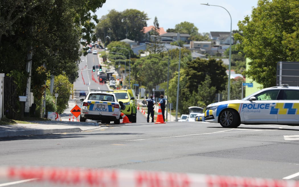 Lights are out at Birkenhead Ave intersection with Onewa Road and traffic is being manually directed by a person in a high-vis vest while armed police guard a cordon.