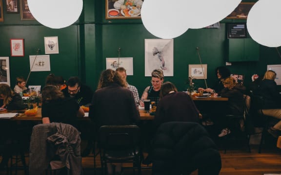 A group of people sit at long tables sketching. They're inside a green-walled cafe strung with big, circular lampshades.