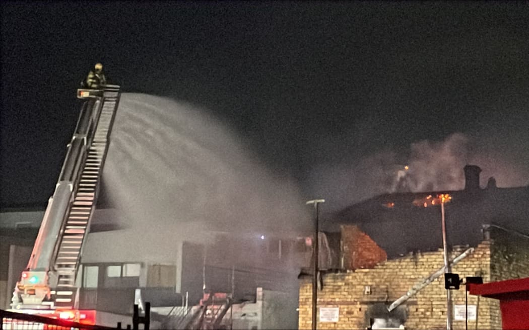 Flames could be seen over the top of a building in Onehunga Mall Road, as firefighters work from ladders, on Sunday night, 25 January, 2026.