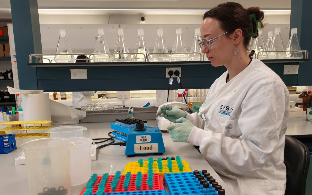 A woman in a white lab coat wearing lab goggles and rubber gloves holds a small sample tube next to a machine. Three brightly coloured trays of other small tubes sit on the lab bench next to her, and a row of conical flasks sits on the shelf behind her.