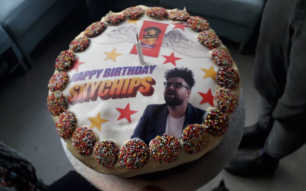 A cake with a photo of Luke Rowell, Wordart text which reads "Happy Birthday Skychips", and a packet of cornchips with angel wings.