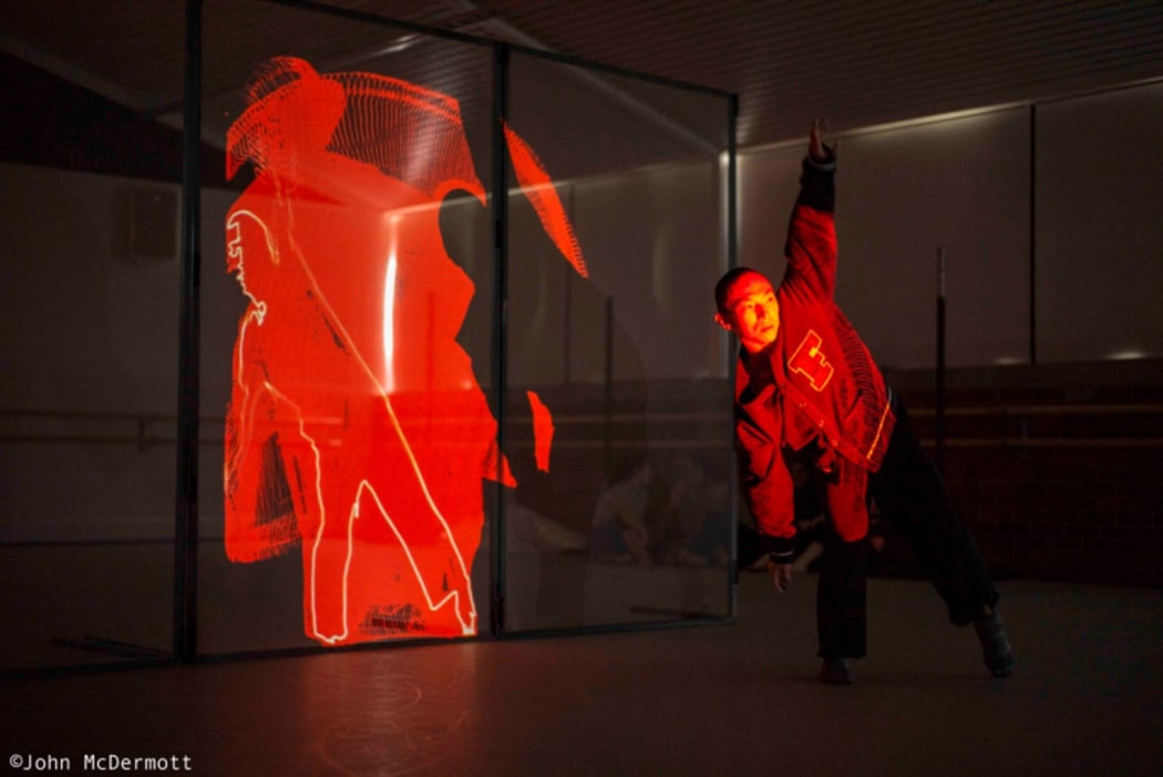 A production still from In Transit showing a performer dancing in front of a clear screen on which his image is being projected.