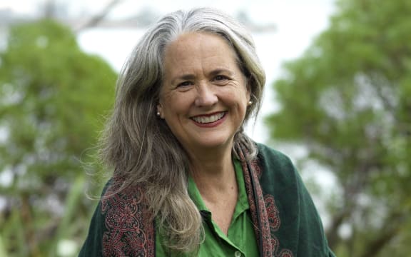 Wendyl smiles at the camera. She is outside in a clearing surrounded by lush bush. The sea is in the distance. It is a bright, pale day.