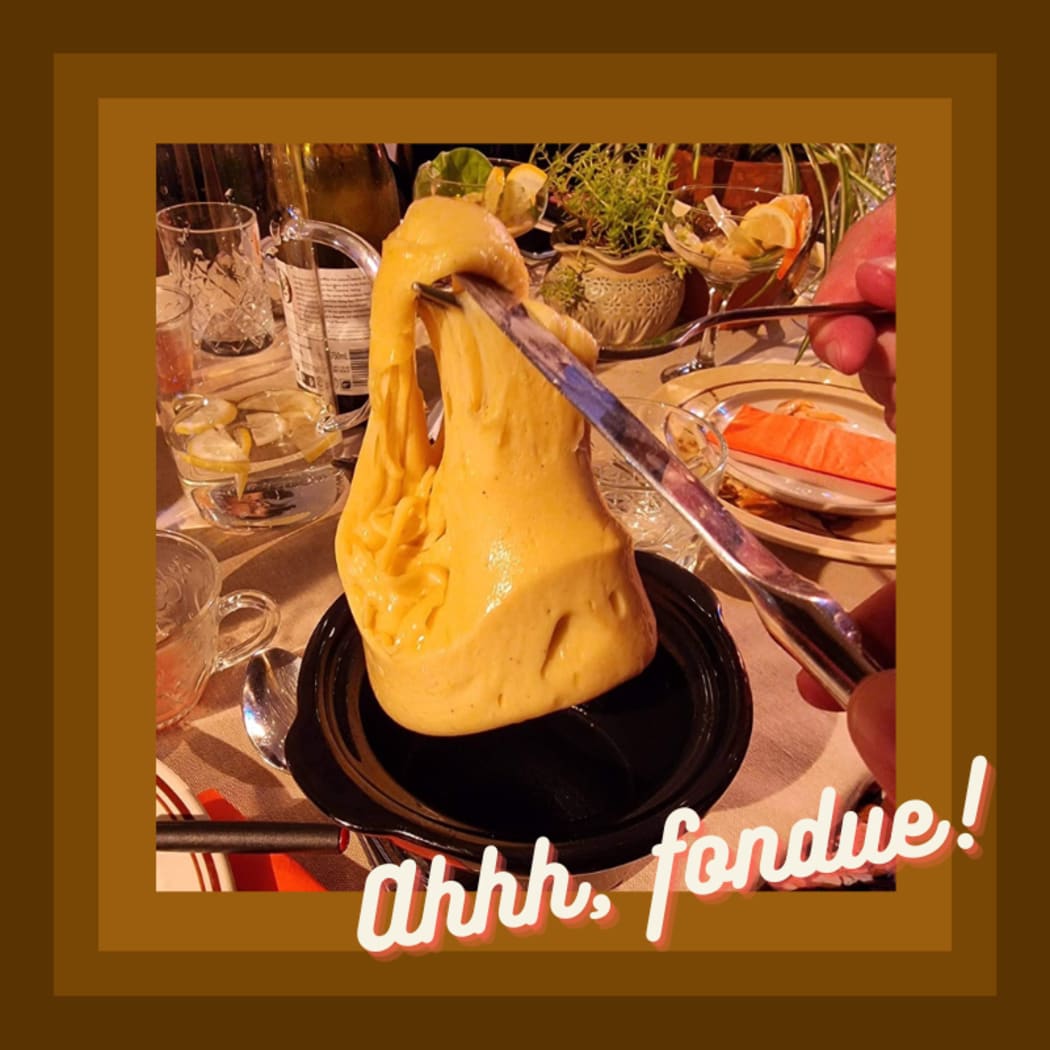 A close up of a greta glob of melted cheese being held up by a knife above a plate. This is another one of the dishes being served at the dinner party. In the backgroudn you can see other dishes and glassware. teh image is bordered by brown 70s tones and captioned witht he words ''Ahh fondue!''
