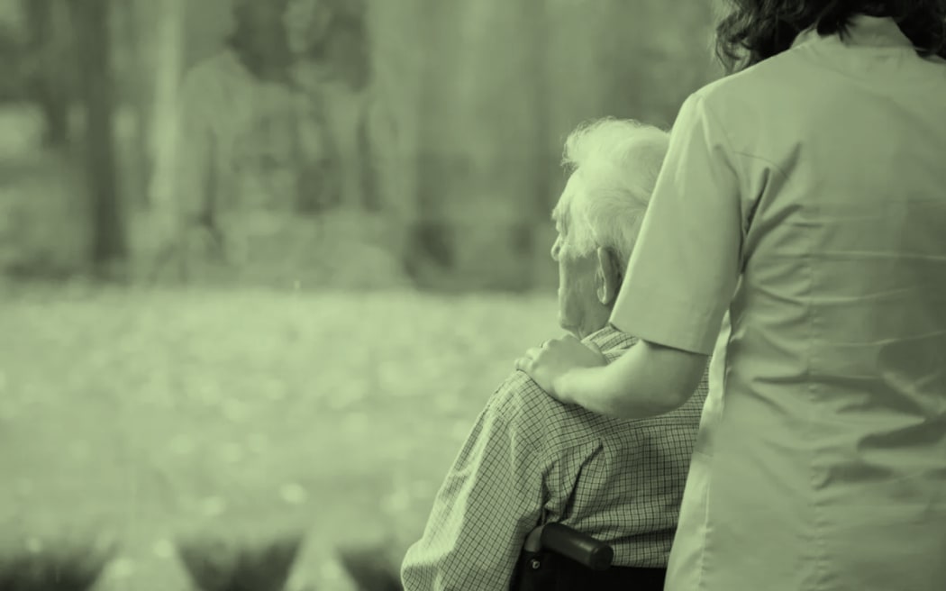 The image shows a residential care nurse standing behind an elderly man sitting in a wheelchair. The image has a light green wash across it.