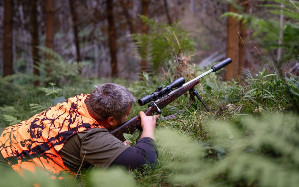 Jasper Osborne is wearing an orange high vis hunting vest. He is lying on the forest floor behind a hunting rifle. The rifle is pointing at something out of frame.