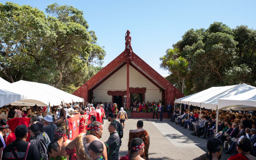A group of wāhine Māori from Te Tai Tokerau stand on the marae atea with their backs turned on the Government in protest during Waitangi commemorations 2025. They are draped in homemade red woollen blankets hand-stitched with Tino Rangatiratanga and He Whakaputanga flags.