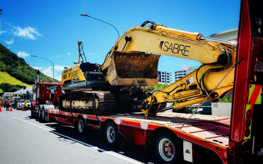 A large digger is brought in to the landslide at Mt , this time with a police escort car guiding it through a residential street
