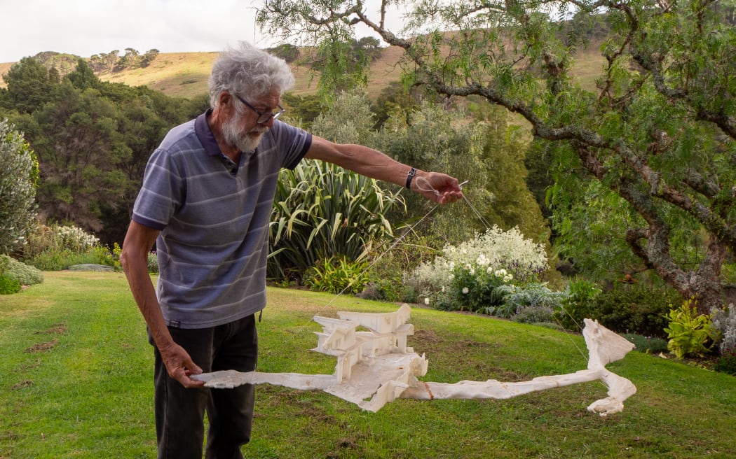 An older man with glasses, standing outside, holds up a 3D-printed model of a house sitting atop a large lava tunnel.