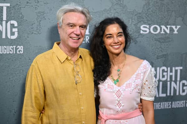 David Byrne, with white hair and wearing a yellow shirt,  stands next to Mala Gaonkar, a dark-haired woman in a pink dress.