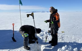 Two people stand next to a rectangular hole cut into the sea ice. The man on the left is wearing all black and he is bending over to insert a long coring drill into the hole. There is a spade and flag on the far left. On the right of the hole, a woman with a short blonde bob stands, holding a rod attached to cables. She is wearing an Antarctica New Zealand black-and-orange jacket. In the background, a flat expanse of white ice extends to low rocky, snow-strewn hills on the horizon, beneath a blue sky with a smear of cloud.