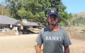 A man wearing a grey tshirt with "BANKS" written on it in white letters and a navy cap with a colourful logo on it looks at the camera. He is standing in front of a house's front yard, which is covered in silt and mud shoveled away into large piles.