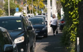 Pedestrians try to use the footpath in Auckland where cars are parking over the kerb.