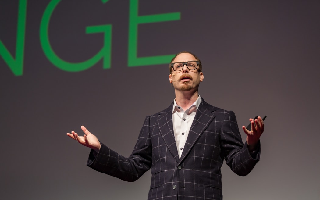Man in dress suit and glasses gives a presentation, extending their arms out, in front of a giant projector screen.