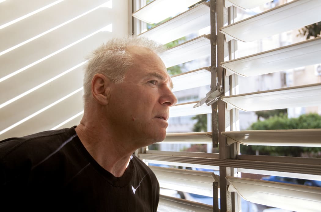 A man in his 60s looks out the window through open blinds. The man is wearing a black t-shirt. His hair gray. Head turned in profile. Glance tense. He looks at what's going on outside the window. Close-up shot.