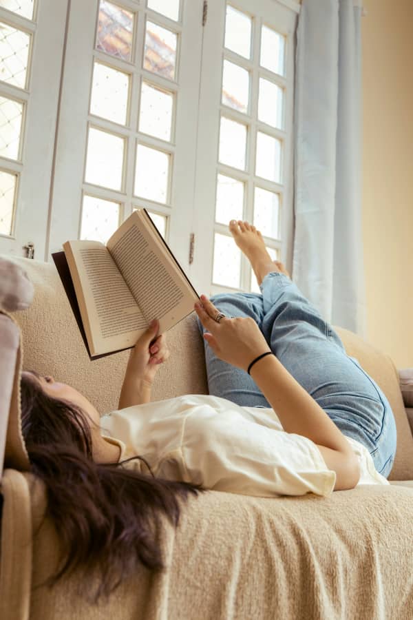 A dark-hared woman lies on the couch reading with her bare feet up.