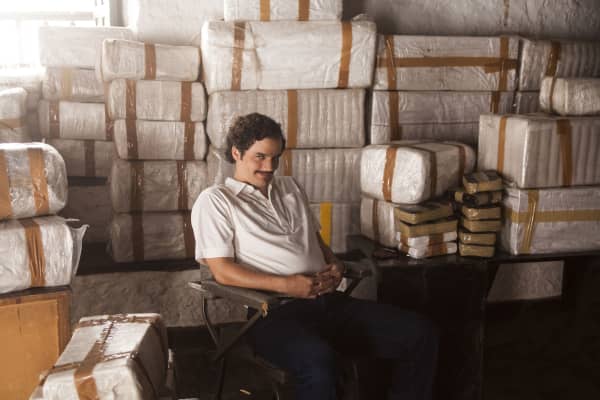 Actor Wagner Moura smiles smugly and wears a white short-sleeve shirt as he sits in front of big packages of a white substance which appears to be cocaine.