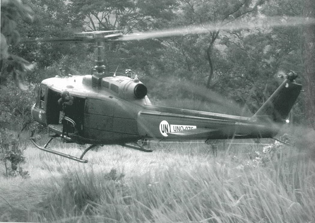 An RNZAF Iroquois hovering just above the ground as it waits to pick up a patrol in the Tilomar area. The door gunner, LAC Daryl Fell, is checking the tail rotor clearance; March 2000.