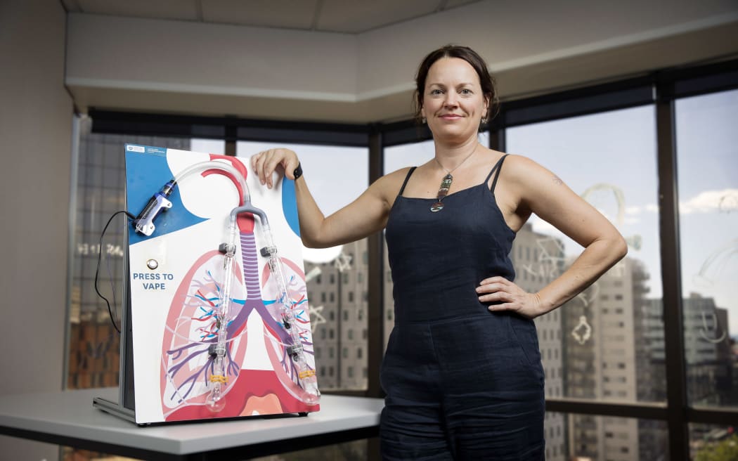 Kelly stands beside an outreach model of how vaping works. There's an image of the lungs with two tubes and a vape attached to a pipe with a button labelled 'press to vape. Kelly is wearing a black string top and black pants, with her hair pulled back, and one hand resting on the outreach device.