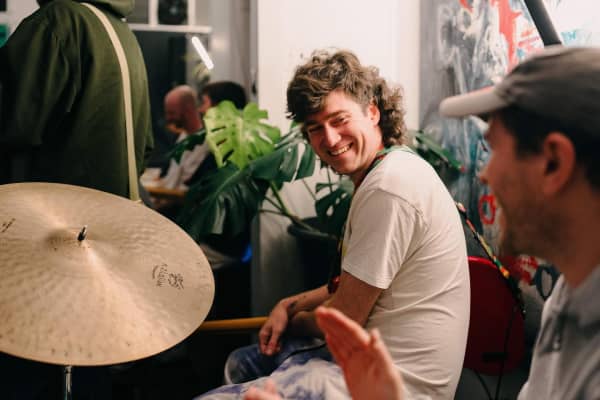 Joshua Worthington-Church is a simling, curly haired man wearing a white t-shirt and a guitar around his neck.