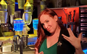 Erin stands in front of a sci-fi set. She is wearing a Star Trek uniform. She smiles and performs the Klingon peace sign for "live long and prosper".