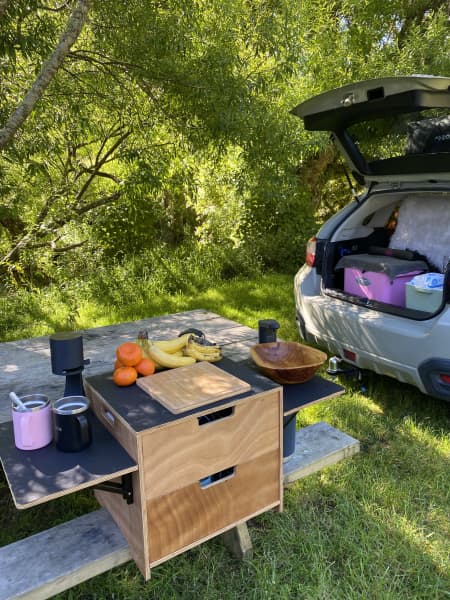 A car boot open outdoors in nature with packed luggage and a bench in the park with fruits, mug and chopping board sitting on top.