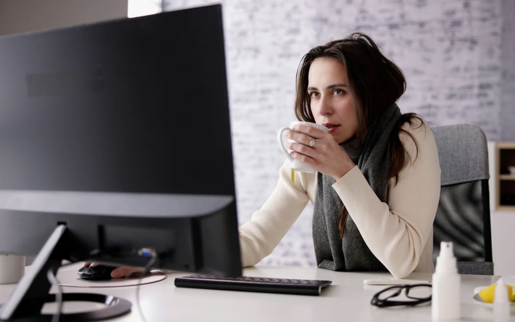 A woman wearing a scarf and knit sweater holds a warm cup of tea at her office desk.