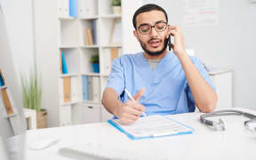 Portrait of young Middle-Eastern doctor wearing glasses sitting at desk in office and speaking by smartphone, filling in patients forms