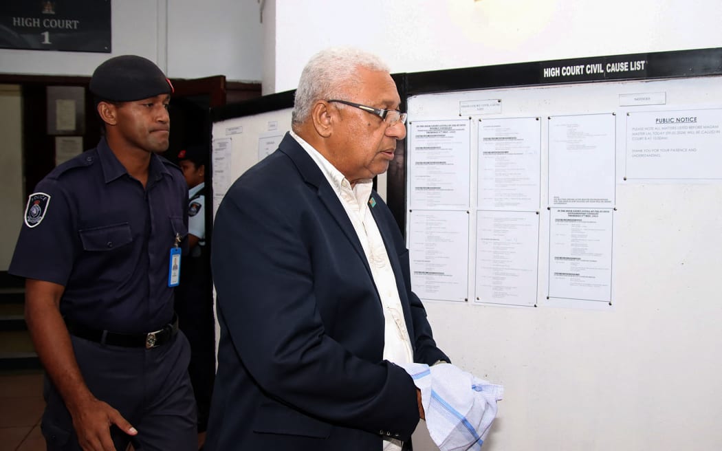 El ex Primer Ministro de Fiji, Frank Bainimarama, esposado, abandona el edificio del tribunal tras recibir su sentencia en Suva el 9 de mayo de 2024. Bainimarama fue condenado el 9 de mayo a un año de prisión por pervertir el curso de la justicia, y un juez concluyó que utilizó su influencia política para cerrar una investigación policial.  (Foto de Kunal KESHNEEL/AFP)