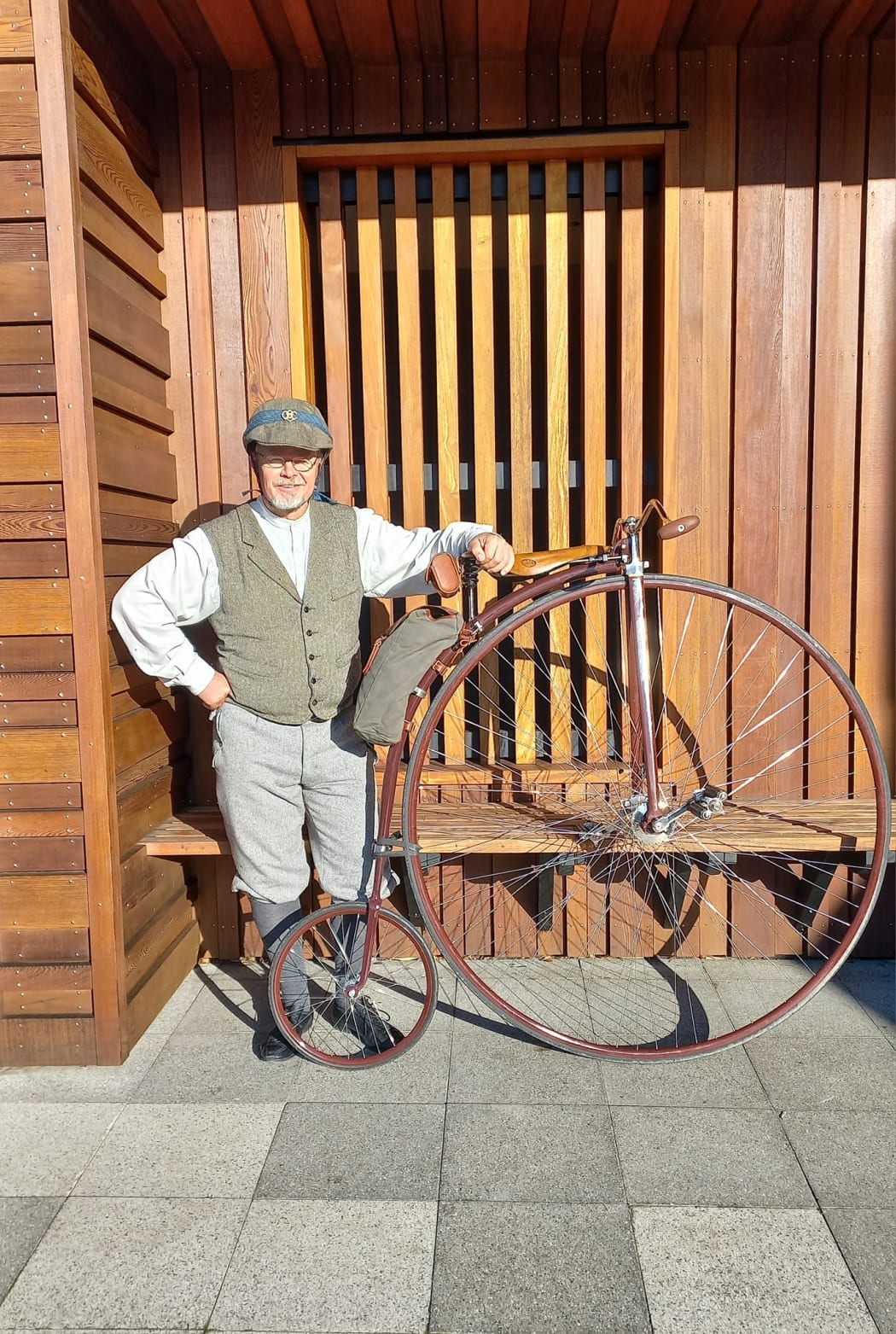 A man dressing in Victorian era clothing stands next to his Penny Farthing, with a wooden lined wall in the background.