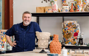 Glenn stands by a table covered in ceramics. He smiles.