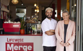 Jo smiles at the camera. She is standing outside a cafe, wearing a pink coat.