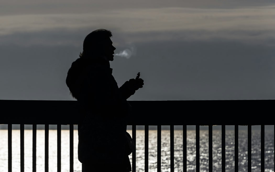 A black-and-white image of a silhouetted person vaping in front of a fence adjacent to a body of water.